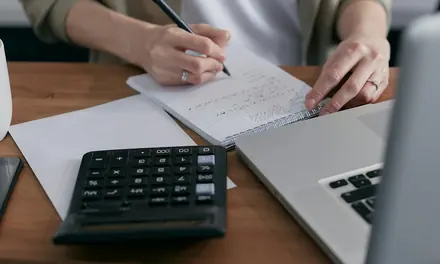 Person writing in a notebook beside a calculator and a laptop