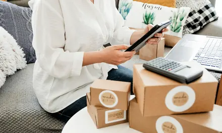 Online seller using a phone and a calculator beside cardboard shipping boxes
