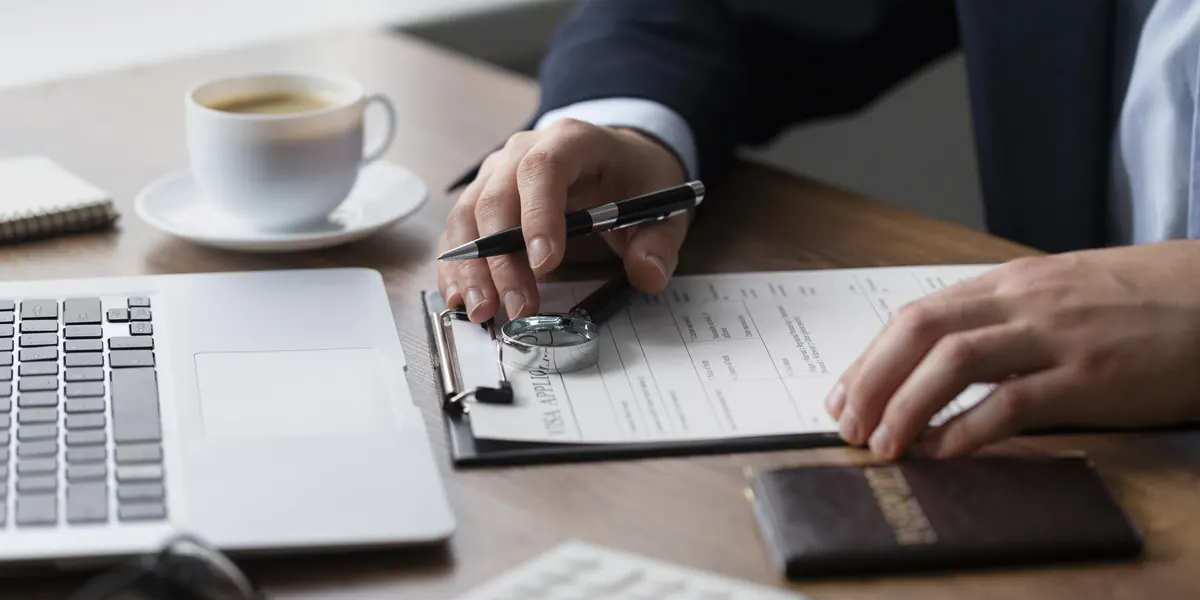 Person signing a form on a clipboard next to a passport, a coffee cup and a laptop