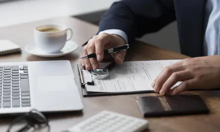 Person signing a form on a clipboard next to a passport, a coffee cup and a laptop