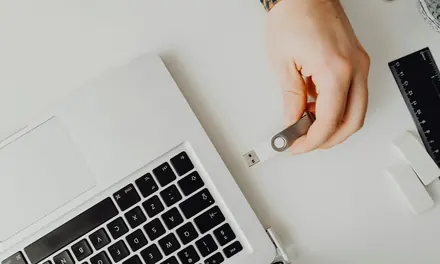 Hand holding a USB flash drive next to a laptop on a white desk